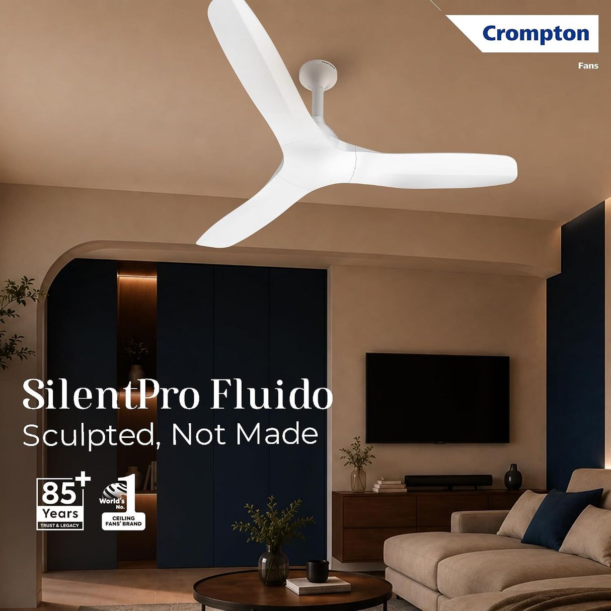 A white Crompton SilentPro Fluido ceiling fan shown installed in a modern living room with beige walls, dark navy cabinets, a sofa, and a TV. Text on the image reads 'Crompton Fans', 'SilentPro Fluido', and 'Sculpted, Not Made', with feature badges in the lower left.