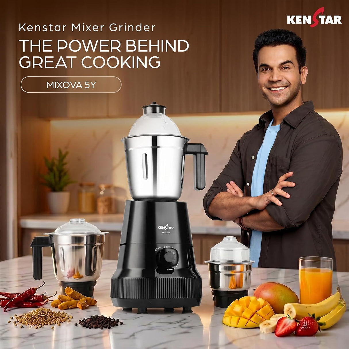 Kenstar Mixer Grinder Mixova 5Y in a kitchen setting with a smiling man in a brown shirt and blue undershirt standing next to it. The main product is a black and stainless steel mixer grinder, with two smaller jars and various fruits and spices arranged on a marble countertop.