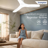 A Crompton ceiling fan with white aerodynamic blades installed in a modern living room, shown above a beige sofa with a child sitting on the couch. The image features promotional text and Crompton branding.