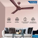 A Crompton ceiling fan in a bronze finish installed in a room with a light pink wall, a white sofa with decorative pillows, and a glass coffee table. The image includes feature callouts for air delivery, speed, star rating, and wattage, along with a Crompton Fans logo in the top right.