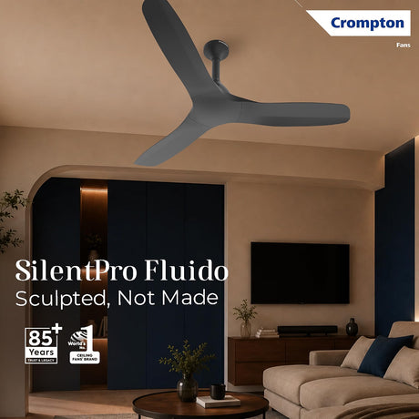 A Crompton SilentPro Fluido ceiling fan in a modern living room setting. The fan has three sculpted blades in a dark gray color.