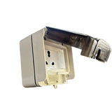 A white plastic wall outlet cover with a transparent protective flap open, resting on a retail display box in a warehouse or store setting with shelving and boxes in the background.