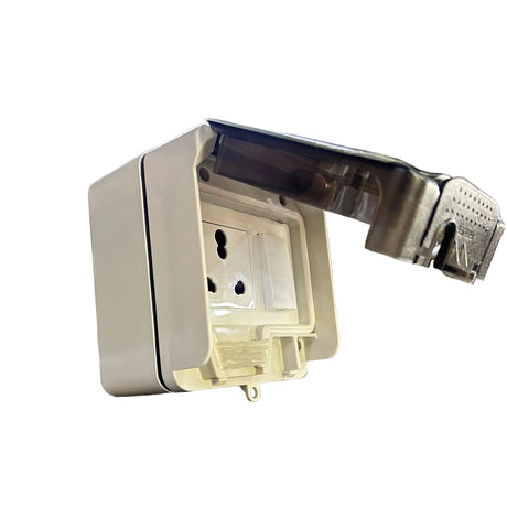A white plastic wall outlet cover with a transparent protective flap open, resting on a retail display box in a warehouse or store setting with shelving and boxes in the background.