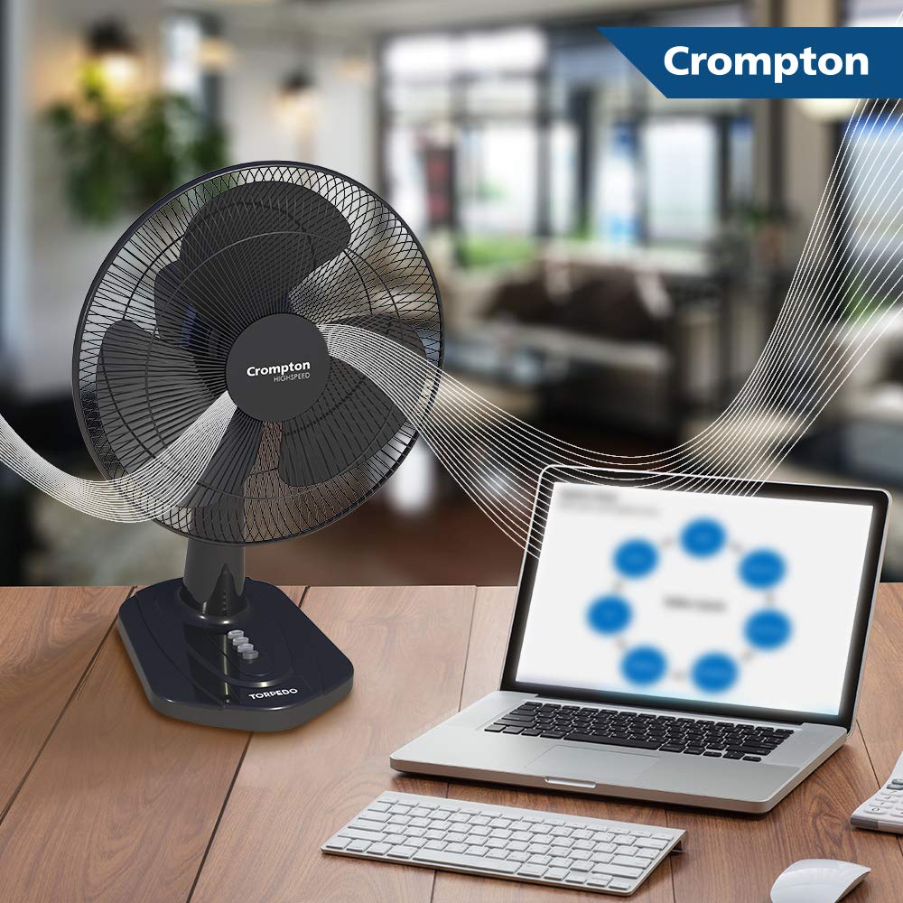 A Crompton desktop fan is shown on a wooden desk next to a laptop and keyboard, with illustrated airflow lines extending from the fan. The background is a blurred indoor room, and a Crompton logo banner appears in the top right corner.