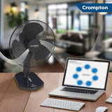 A Crompton desktop fan is shown on a wooden desk next to a laptop and keyboard, with illustrated airflow lines extending from the fan. The background is a blurred indoor room, and a Crompton logo banner appears in the top right corner.