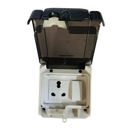 A waterproof outdoor wall outlet cover with a transparent dark gray waterproof cap and a white plastic outlet insert, shown open on a scratched white surface.