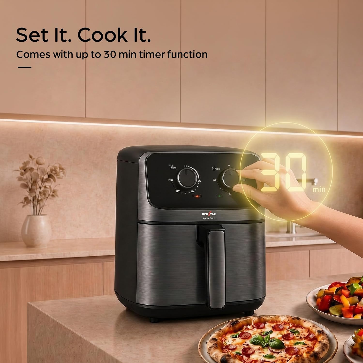 A black and stainless steel air fryer sits on a kitchen counter with a hand adjusting its digital timer display, which shows 30 minutes. The background is a warm beige kitchen setting with food items like pizza and salad visible on the counter, and promotional text in the upper left corner.