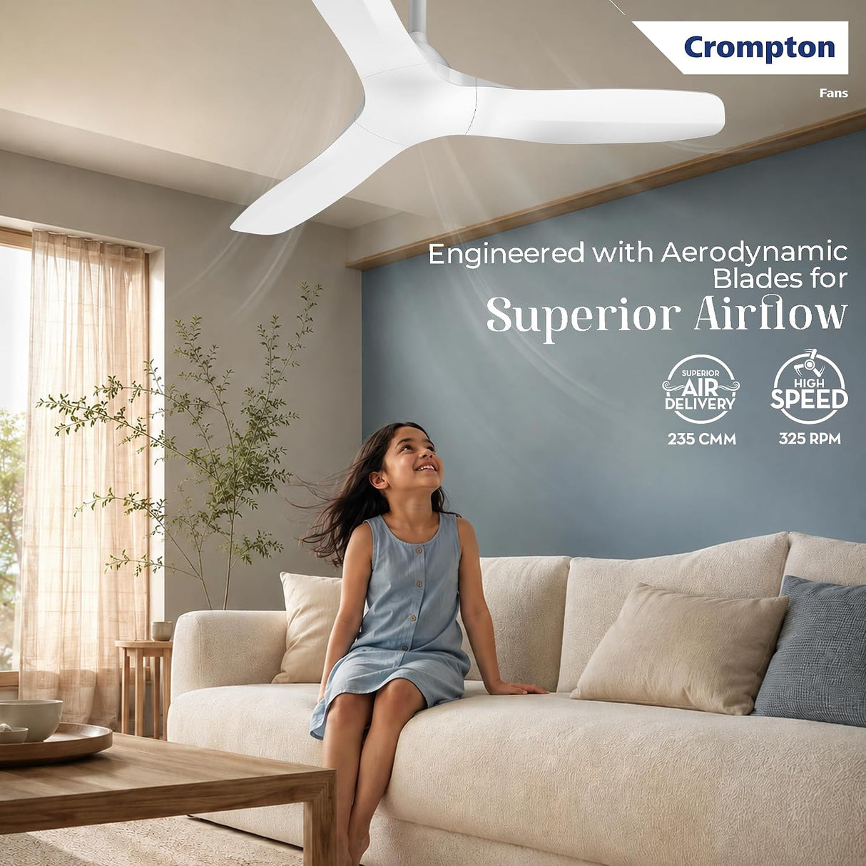A woman sits on a cream sofa in a modern living room while a white Crompton ceiling fan blows air toward her. The room has a blue accent wall with product text and Crompton branding.