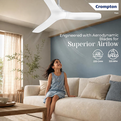 A woman sits on a cream sofa in a modern living room while a white Crompton ceiling fan blows air toward her. The room has a blue accent wall with product text and Crompton branding.