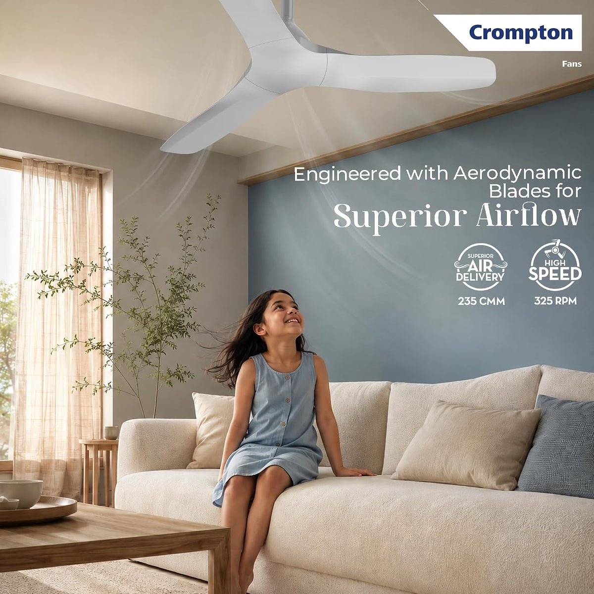 A white Crompton ceiling fan shown in a modern living room above a beige sofa, with a woman seated on the couch. The image includes promotional text and icons for aerodynamic blades, 235 cm airflow, and 325 RPM speed.
