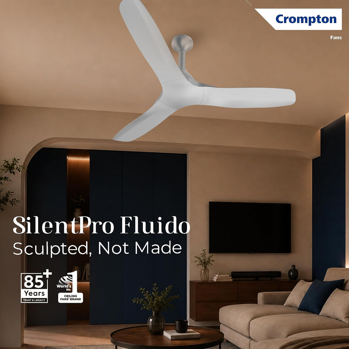 A white Crompton SilentPro Fluido ceiling fan installed in a modern living room with beige walls, a sofa, and a TV. The image includes promotional text and the Crompton Fans logo.