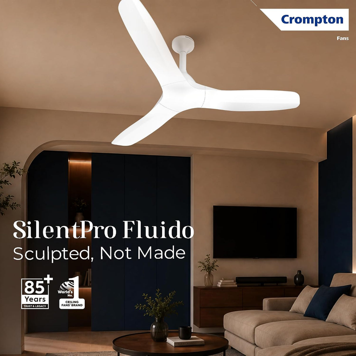 A modern three-blade ceiling fan by Crompton shown installed in a stylish living room. The fan has a sculpted white body and is mounted on a beige ceiling.