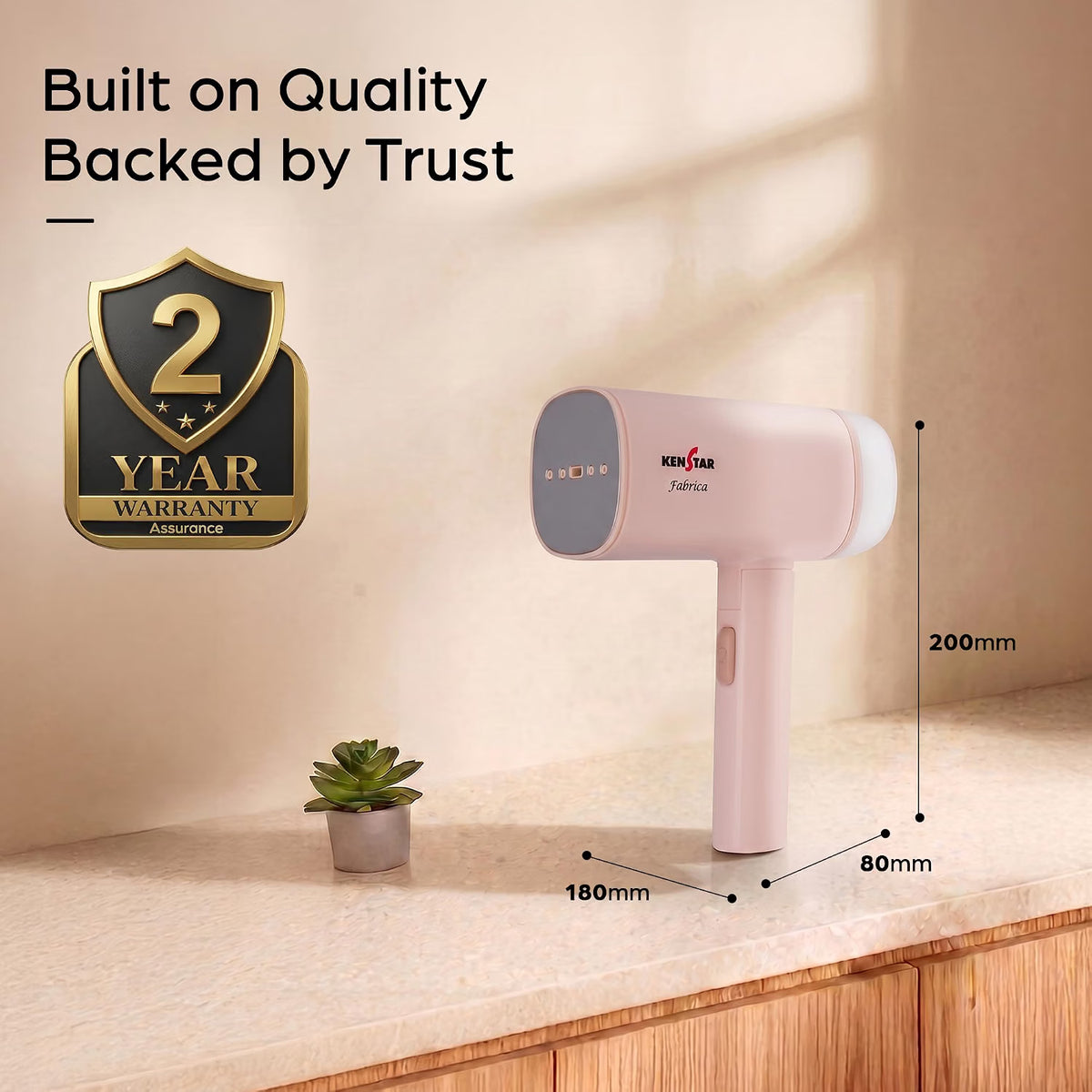 Pink handheld vacuum cleaner shown on a countertop with dimension labels (180mm width, 80mm depth, 200mm height) and a 2-year warranty badge. The background is a warm beige wall with soft shadows.
